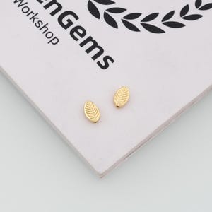 May include: Two small, gold-colored metal beads shaped like leaves with a textured vein pattern. The beads are positioned on a white card featuring the black printed text "enGems Workshop" and a black laurel wreath graphic at the top corner.