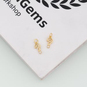 May include: Two gold-toned question mark charms, each featuring a small clear stone at the base of the punctuation mark. The charms include a circular jump ring at the top for attachment, showcasing a polished metallic finish and a delicate design.