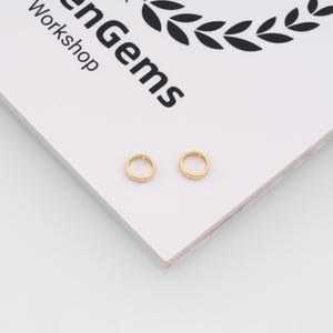 May include: Two small, circular gold-toned metal rings with a smooth finish, each featuring a single tiny embedded clear stone. The rings are positioned on a white surface with the black text "enGems Workshop" and a laurel wreath logo visible.