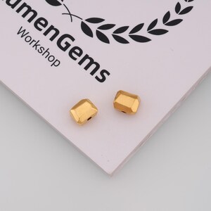 May include: Two small, gold-coloured, faceted beads with a rectangular shape and a central hole, resting on a white card featuring the black printed text "LumenGems Workshop" and a partial laurel wreath graphic at the top edge of the card.