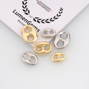 May include: An assortment of gold and silver-colored bean-shaped jewelry components. The components have two holes and are arranged on a white surface near a paper with the text "Lumen Gems Workshop".