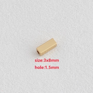 Puede incluir: Un pequeño espaciador rectangular de metal dorado con un agujero. El texto en la imagen dice "size:3x8mm hole:1.5mm".