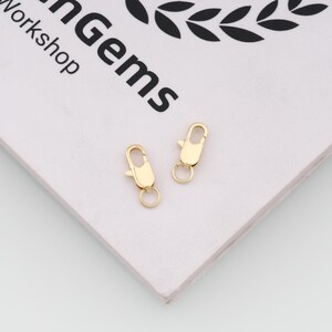 May include: Two small gold-toned lobster claw clasps with attached jump rings. The metal fasteners feature a spring-loaded mechanism and a smooth, polished finish. They are positioned on a white surface with the text nGems Workshop visible.