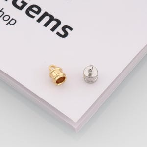 May include: Two small metal jewelry components. One is gold-colored with a loop and a cylindrical shape. The other is silver-colored, also cylindrical, with a small post on top. The word "gems" is visible in the background.