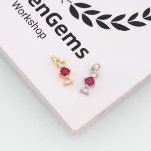 Pode incluir: Dois pingentes em forma de doce com uma pedra vermelha redonda central ladeada por duas pedras rosas triangulares menores. Um pingente é dourado e o outro prateado, ambos com um pequeno anel no topo. O texto "enGems Workshop" é visível.