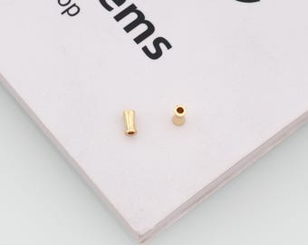 May include: Two small, cylindrical gold-toned beads with a smooth, polished finish. Each bead features a central hole running through its length, designed for threading. The beads are positioned on a white surface with partial black text.