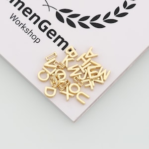 May include: A collection of small, gold-colored metal alphabet letter charms scattered on a white surface featuring the black printed text "menGem Workshop" and a laurel wreath graphic. Each individual letter charm has a polished finish.