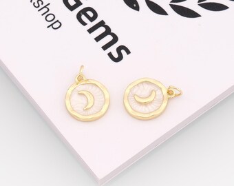 May include: Two gold-colored round pendants with a white crescent moon design. The pendants have a gold border and a small loop at the top for attaching to a chain or other jewelry component. The background is a white and pink surface.