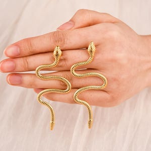 May include: A pair of gold-coloured snake earrings. The earrings are shaped like a snake with a detailed scale pattern. The snake's head is at the top, and the body curves down. The earrings are displayed on a hand.