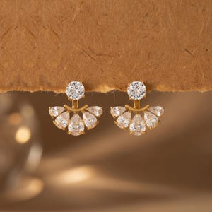 May include: A pair of gold-toned earrings with a post and a large, round, clear stone at the top. Below, a fan-shaped design features five pear-shaped, clear stones. The earrings are set against a brown background.