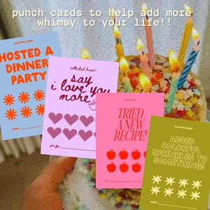 May include: A selection of colourful punch cards with phrases such as "Hosted a Dinner Party" and "Say I love you more." The cards are in shades of blue, pink, and green, with orange and red accents. The cards are next to a cake with lit candles.