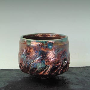 May include: A handcrafted ceramic bowl with a textured surface and a distinctive glaze. The bowl displays a mix of colours, including copper, teal, and brown. The bowl's exterior has a carved pattern. The bowl is suitable for serving or display.