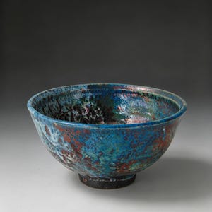 May include: A ceramic bowl featuring a vibrant, multi-coloured glaze with deep blue, turquoise, and reddish-brown tones. The surface has a textured, mottled appearance with a glossy finish, and the bowl rests on a dark, circular base.