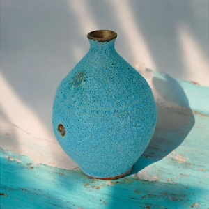 Può includere: Un vaso in ceramica turchese con superficie strutturata e corpo arrotondato. Il vaso ha un collo stretto con bordo dorato e base marrone. Il vaso è alto circa 20 cm.