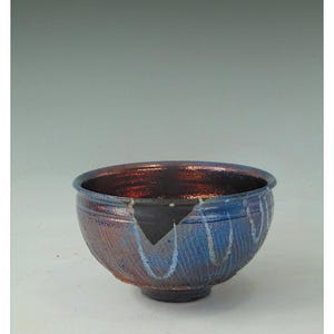 Könnte beinhalten: Eine handgefertigte Keramikschale mit einem einzigartigen Design. Die Schale weist eine Kombination aus blauen und rotbraunen Farben auf, mit einer strukturierten Außenseite und einer glatten Innenseite. Die Schale hat einen Bruch, der ihren Charakter unterstreicht.