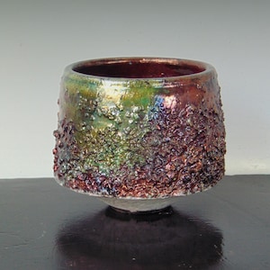 May include: A handcrafted ceramic bowl with a textured, iridescent surface. The bowl features a rich colour palette of green, purple, and brown, with a dark interior. The bowl is approximately 13 cm in diameter and 10 cm tall.