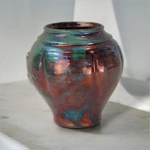 May include: A ceramic vase with a rounded body and a flared rim. The vase is a mix of colours, including deep red, teal, and green, creating a marbled effect. The surface has a glossy finish, reflecting light. This decorative vase is suitable for home decor.