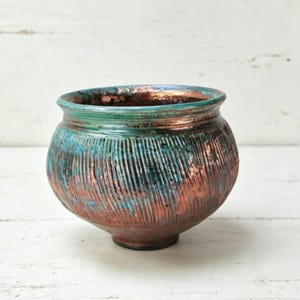 May include: A round ceramic bowl featuring a textured exterior with vertical ridges. The surface displays a metallic copper finish blended with vibrant teal and turquoise accents, creating a multi-toned appearance across the rounded body.