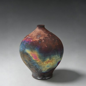May include: A round ceramic vase with a narrow neck and a wide body. The surface features a dark, mottled finish with iridescent patches of blue, yellow, and pink, contrasted by areas of deep brown and black, with subtle horizontal ring textures.