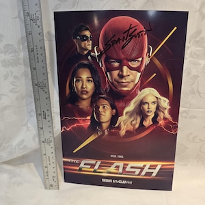 May include: A promotional poster for the TV show "The Flash" featuring several cast members. The poster has a red and gold colour scheme with the show's title prominently displayed. The poster is approximately 30 cm tall.