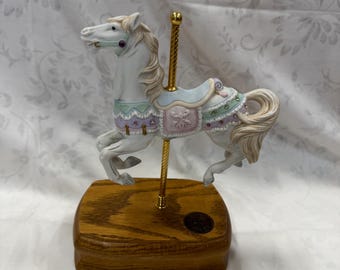 Puede incluir: Un caballo de carrusel de porcelana blanca con una silla de montar rosa, azul y verde y un poste dorado. El caballo está sobre una base de madera con una etiqueta dorada.