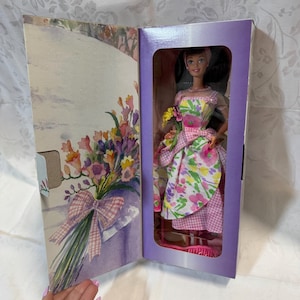 Peut inclure: Une poupée Barbie vêtue d'une robe en vichy rose et blanche à imprimé floral. La poupée tient un bouquet de fleurs et se tient dans une boîte violette sur fond blanc.