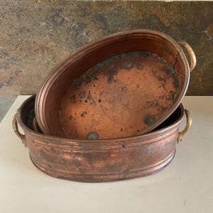 May include: Two oval copper basins with a weathered patina and visible green oxidation spots. Each basin features two curved brass handles on the sides. The metal surfaces show signs of age and use, with a warm metallic tone and dark marks.