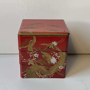 May include: Red, stacked, lacquered box with gold painted crane and floral design. The box has a rectangular shape with a lid and two compartments. The crane and floral design is painted on the front and sides of the box. The box is likely used for storage or display.