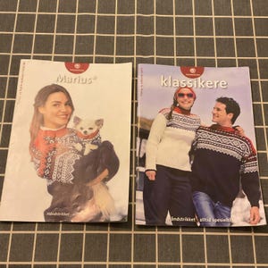 Puede incluir: Dos revistas con personas que llevan suéteres estampados. La revista de la izquierda se titula "Marius" y muestra a una mujer sosteniendo un perro pequeño, ambos con suéteres a juego. La revista de la derecha se titula "klassikere" y muestra a una pareja con suéteres a juego.