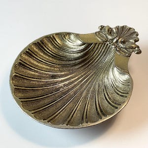 May include: A gold-toned metal dish shaped like a scallop shell. The surface features deep, radiating ridges that extend from the centre to the outer edge. At the top, a decorative, ornate hinge-like detail adds texture to the shell's curved form.