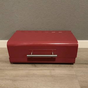 May include: A red bread bin with a curved top and a stainless steel handle. The rectangular box sits on a wooden surface. The bread bin has small ventilation holes along the top edge. The dimensions are approximately 40 cm in length.