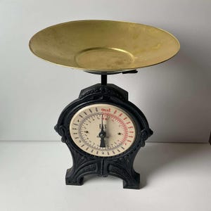 May include: A vintage black and gold kitchen scale. The scale features a circular dial with a needle and measurements in ounces. The weighing pan is gold-coloured, and the base is black with decorative detailing. The scale measures up to 454 grams.