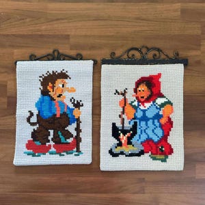 Pode incluir: Duas tapeçarias de parede vintage em ponto cruz. Uma retrata um homem em traje azul e castanho com um bastão, a outra uma mulher com capuz vermelho e vestido azul. Ambas são montadas em cabides de metal decorativos, mostrando arte popular.