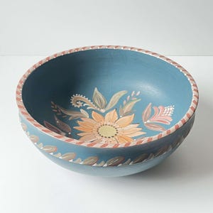 Puede incluir: Un tazón de madera azul con un diseño floral pintado a mano. El tazón presenta un girasol con pétalos naranja y amarillos, hojas verdes y flores rosas.