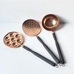 May include: Three copper kitchen utensils with black wooden handles. The utensils include a ladle, a slotted spoon, and a small pan with six indentations.