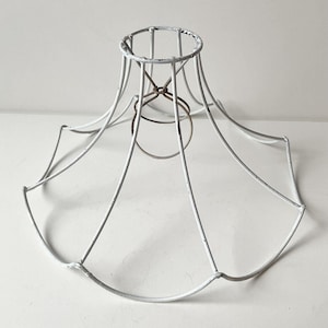 Op de afbeelding: Een witte lampenkapframe in vintage-stijl. Het metalen frame heeft een verweerde afwerking, met een cirkelvormige bovenkant en een uitlopende, geometrische vorm. Het frame heeft een kleine, cirkelvormige ring in het midden voor de bevestiging van de lamp.