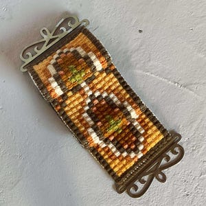 Puede incluir: Un tapiz rectangular vintage con un patrón geométrico en tonos marrón, naranja, amarillo, blanco y verde. El tapiz está enmarcado por un borde metálico con adornos decorativos. El textil es probablemente tejido a mano.