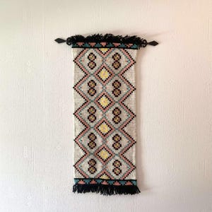 Puede incluir: Un tapiz de pared tejido rectangular con un patrón geométrico en crema, rosa, amarillo y negro. El tapiz tiene una franja negra y está montado con herrajes negros.