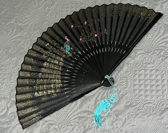 May include: Black folding hand fan with a painted design featuring gold accents and pink and teal details. The fan has a teal tassel attached to the base. The fan is open, displaying the intricate design.