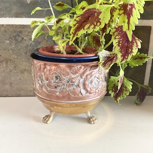 May include: A round copper-toned metal planter featuring an embossed rose pattern around the exterior. The planter has a smooth brass-coloured base with three claw-style feet and contains a dark blue inner pot holding a green and maroon coleus plant.