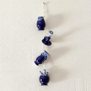 Può includere: Una fila di quattro piccoli vasi in ceramica blu scuro con dettagli bianchi, sospesi a una corda contro un muro bianco strutturato. I vasi hanno forme diverse, tra cui un'urna piccola, un portacandele e due vasi con aperture svasate.