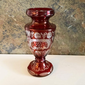 Peut inclure: Un vase en verre rouge rubis avec des motifs floraux et géométriques gravés. Le vase a un corps bulbeux, un bord évasé et une base en piédestal. Les détails gravés sont d'une couleur plus claire, créant un contraste avec le verre rouge.