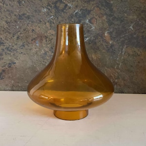 Puede incluir: Un jarrón de vidrio ámbar vintage con cuerpo redondeado y cuello acampanado. El jarrón se asienta sobre una pequeña base circular. El vidrio tiene un acabado brillante, que refleja la luz. Este jarrón decorativo es adecuado para flores o como pieza de arte.