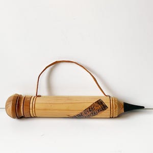 May include: A wooden pencil-shaped object with a brown leather handle. The pencil has a light brown body, a dark brown eraser end, and a black tip. A label with text is affixed to the body.