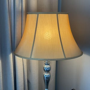 Pode incluir: Uma lâmpada alta e elegante com uma abajur bege e uma base prateada polida. O abajur tem uma forma clássica com uma borda verde clara. A lâmpada está posicionada em frente a uma cortina cinza claro.