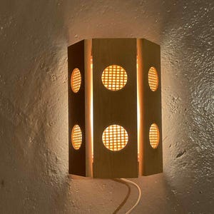 May include: A wooden wall sconce with a geometric design. The sconce has a cylindrical shape with six sides. Each side features a circular cutout with a grid pattern, allowing light to shine through. The sconce is mounted on a textured wall.