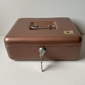May include: A bronze-coloured, rectangular cash box with a textured finish. It has a silver handle, a key lock with a key inserted, and a small logo on the front. The box is designed for secure storage of money and valuables.