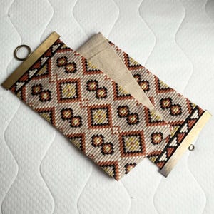 Puede incluir: Textil tejido con un patrón geométrico en crema, naranja, amarillo y negro. Tiene barras metálicas color latón en cada extremo, con un anillo en un extremo. El textil está doblado sobre una tela beige.