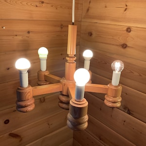 May include: A light-colored wooden chandelier featuring a central vertical post and four horizontal arms, each ending in a cylindrical base with a white socket and a glowing light bulb. The fixture is suspended by a single white cord from above.
