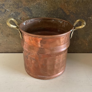 May include: A cylindrical copper bucket featuring a hammered texture and a polished finish. The vessel includes two curved brass handles securely attached to opposite sides of the rim with metal rivets, providing a contrast to the warm copper body.